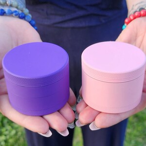 May include: Two metal grinders, one purple and one pink. Both grinders have a textured surface and are four-piece grinders.