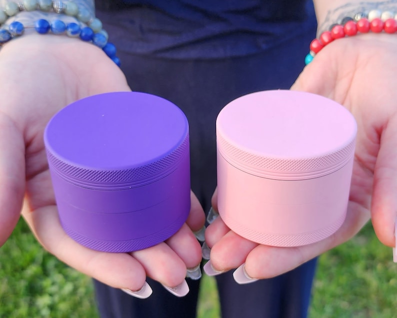 May include: Two metal grinders, one purple and one pink. Both grinders have a textured surface and are four-piece grinders.