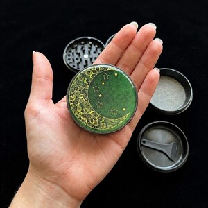 K&ouml;nnte beinhalten: Eine kreisf&ouml;rmige Kr&auml;uterm&uuml;hle mit einem dunkelgr&uuml;nen Deckel, der ein goldenes Mondsichel-Design mit komplizierten Blumenmustern und kleinen Sternumrissen zeigt. Die M&uuml;hle ist in vier schwarze Komponenten zerlegt, inklusive Sieb und kleinem Schaber.