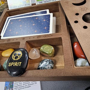 Peut inclure: Un présentoir en bois pour cartes de tarot avec compartiments, contenant un jeu de cartes avec un motif de ciel étoilé et diverses pierres polies. Une pierre noire avec le mot "SPIRIT" est visible. Le présentoir a un couvercle décoratif.