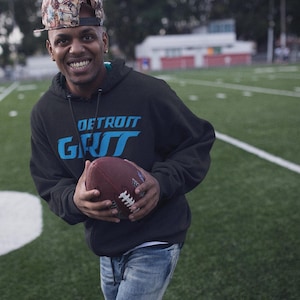 May include: A person wearing a black hoodie with the text "DETROIT GRIT" in blue lettering. The person is holding a brown football.