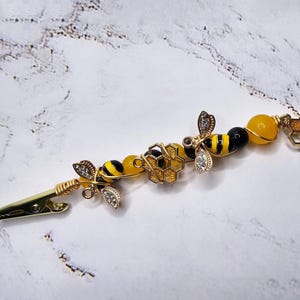 K&ouml;nnte beinhalten: Eine goldene Haarklammer mit einem Bienen- und Wabenmuster. Die Klammer ist mit gelben und schwarzen Perlen, goldfarbenem Draht und einem kleinen Waben-Anh&auml;nger verziert.