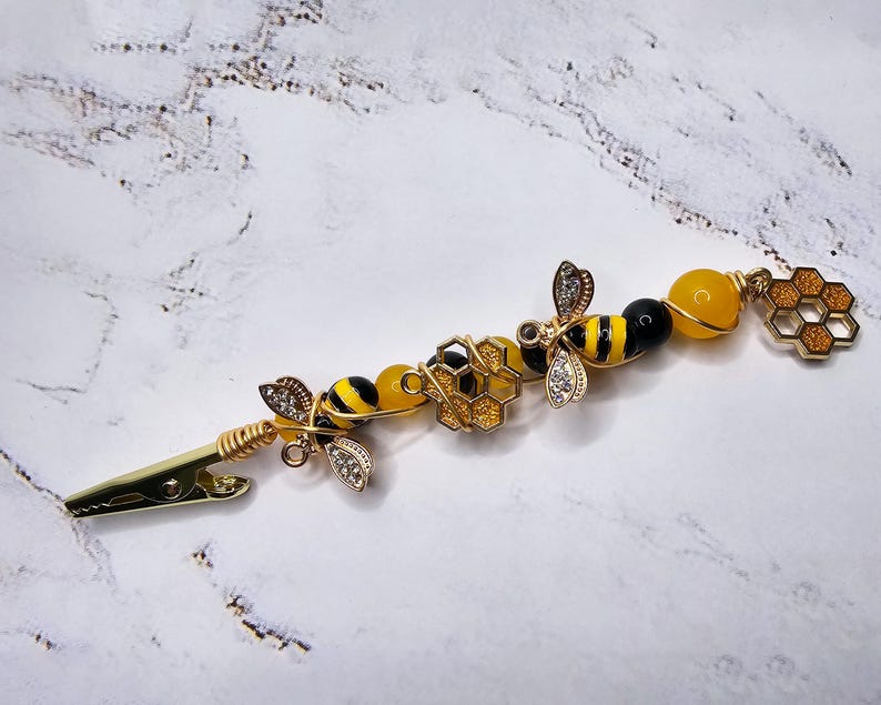 K&ouml;nnte beinhalten: Eine goldene Haarklammer mit einem Bienen- und Wabenmuster. Die Klammer ist mit gelben und schwarzen Perlen, goldfarbenem Draht und einem kleinen Waben-Anh&auml;nger verziert.