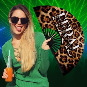 Puede incluir: Un abanico de mano plegable negro con un diseño de estampado de leopardo y la palabra "FERAL" en blanco. El abanico se sostiene sobre un fondo verde. La imagen incluye a una mujer con gafas de sol y un top verde.