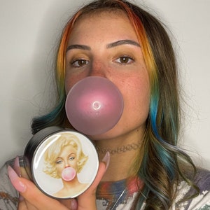 May include: A round, black and silver container with a white lid featuring a portrait of Marilyn Monroe blowing a bubblegum bubble. The container is held in front of a person with rainbow-colored hair and a pink bubblegum bubble.