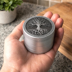 Puede incluir: Un molinillo de hierbas cilíndrico de metal color plata con un diseño de árbol en relieve detallado en la tapa superior. La tapa está rodeada por un borde que contiene símbolos de estilo rúnico y el lateral tiene un patrón de agarre texturizado.