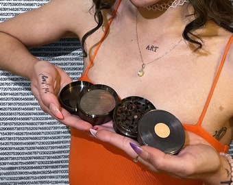 May include: A close-up of a black metal grinder with a record-shaped top. The grinder is in the hand of a person wearing an orange shirt. The person has a tattoo on their finger that says "Bad-m-f". The person also has a necklace with a small diamond pendant and the word "ART" on it.