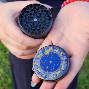 Könnte beinhalten: Ein zweiteiliger Grinder in Schwarz und Blau. Der obere Teil ist schwarz mit einem Gitter aus rautenförmigen Löchern. Der untere Teil ist blau mit einem goldenen Tierkreisdesign. Der Grinder wird in offenen Händen gehalten.