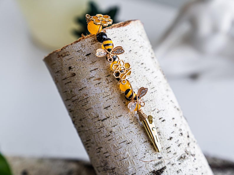 K&ouml;nnte beinhalten: Eine goldene Bienen-Haarklammer mit gelben und schwarzen Perlen, einem Waben-Anh&auml;nger und einem Bienen-Anh&auml;nger. Die Klammer befindet sich auf einem wei&szlig;en Birkenholz-Hintergrund.