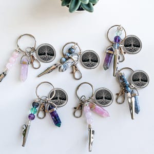 Puede incluir: Seis llaveros plateados con un diseño de árbol de la vida, cada uno con un dije de cristal diferente. Los cristales incluyen amatista, cuarzo rosa, fluorita y apatita azul.