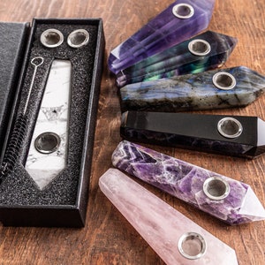 May include: A set of seven natural gemstone smoking pipes in various colors, including purple, green, black, and pink. The pipes are in the shape of points and are arranged in a semi-circle on a wooden surface. The pipes come in a black box with the words "Natural gemstone smoking pipes" printed on the front.
