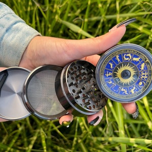 Könnte beinhalten: Ein zerlegter Kräutermühle mit einem Sternzeichen-Design. Der Grinder ist silber und schwarz, mit einem blauen und goldenen Sternzeichenrad auf der Oberseite. Der Grinder wird in einer Hand vor einem Hintergrund aus grünem Gras gehalten.