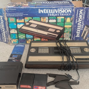 Puede incluir: Una consola de videojuegos Intellivision con su caja original, mandos y cartuchos de juego. La consola es negra y beige con acabado de vetas de madera. La caja es azul con el logotipo de Intellivision y gráficos de juegos.