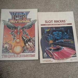 Puede incluir: Dos manuales de instrucciones de juegos Atari antiguos. "Yars' Revenge" presenta una ilustración de insecto robótico sobre un fondo rojo y naranja. "Slot Racers" tiene una ilustración de coche de carreras rojo sobre fondo azul. Ambos manuales están sobre una superficie gris.