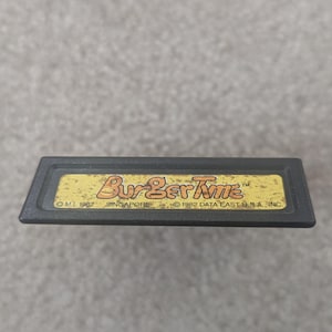 Könnte beinhalten: Eine schwarze, rechteckige Videospielkassette mit dem Titel "BurgerTime" in gelben und orangefarbenen Buchstaben. Die Kassette enthält auch den Text "© M.I. 1982 SINGAPORE © 1982 DATA EAST U.S.A. INC."