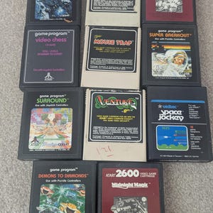 Puede incluir: Una colección de cartuchos de videojuegos Atari 2600. Los títulos incluyen "Asteroids", "Donkey Kong", "Super Breakout", "Space Jockey" y "Demons to Diamonds". Los cartuchos presentan etiquetas coloridas con gráficos y texto.