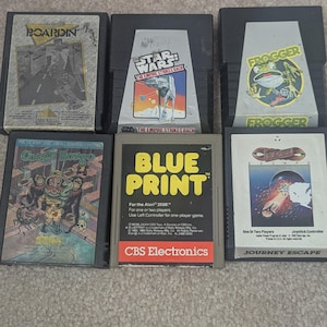 Puede incluir: Una colección de cartuchos de videojuegos Atari 2600 antiguos. Los títulos incluyen "Boarding", "Star Wars: The Empire Strikes Back", "Frogger", "Congo Bongo", "Blueprint" y "Journey Escape". Los cartuchos presentan ilustraciones de juegos coloridas.