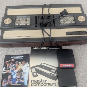 May include: A vintage Mattel Intellivision video game console, with controllers, power cord, and game cartridges. Includes manuals and cartridges labelled "Kool-Aid Man" and "B-17 Bomber". The console is black and beige.