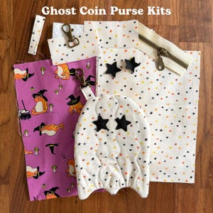 May include: Ghost Coin Purse Kits. A sewing kit featuring a purple fabric with orange frogs in black witch hats, a white fabric with small multicoloured stars, a quilted ghost-shaped piece with black star eyes, metal zips, and lobster clasps.