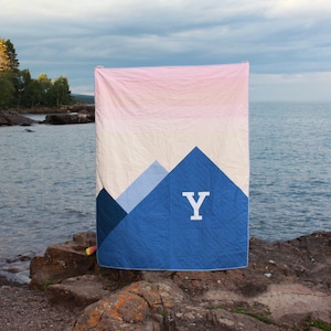 May include: A blue and pink quilt with a mountain design and the letter "Y" in white in the center. The quilt is hanging on a clothesline against a backdrop of a lake and trees.