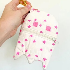 Może przedstawiać: Biała portmonetka w kształcie ducha z zapięciem na zamek błyskawiczny i złotym zapięciem. Portmonetka jest ozdobiona różowymi wzorami gwiazd i różowymi kwadratowymi oczami. U góry znajduje się napis "GHOST COIN PURSE".