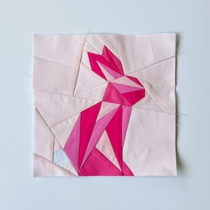 May include: A pink geometric bunny quilt block with a white background. The bunny is made of various shades of pink fabric and is pieced together in a geometric pattern.