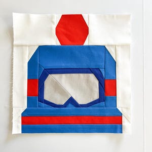 May include: A blue and red patchwork quilt block with a geometric design. The block features a red hexagon in the center, surrounded by a blue square with a white diamond shape inside. The blue square is bordered by red strips.