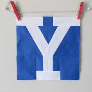 Puede incluir: Un bloque de tela azul y blanco con una forma de 'Y' blanca en el centro. El bloque está colgado de una cuerda con pinzas para la ropa.