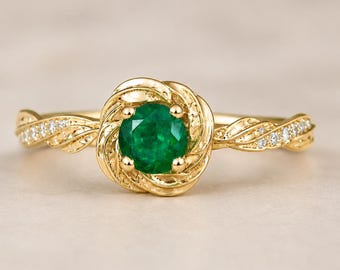 Könnte beinhalten: Ein goldener Ring mit einem runden, leuchtend grünen Smaragd im Zentrum. Der Ring hat eine blumenförmige Fassung und ein verdrehtes Band mit kleinen, funkelnden Akzentsteinen. Der Ring ist vor einem neutralen Hintergrund platziert.