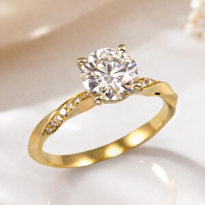 Puede incluir: Un anillo de compromiso de oro con una piedra preciosa grande, redonda y transparente. La banda tiene un diseño retorcido con pequeñas piedras de acento. El anillo está sobre un fondo blanco suave con acentos florales.
