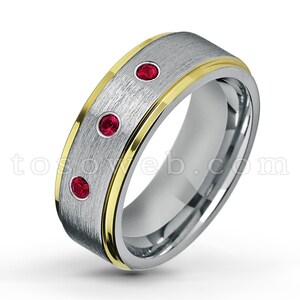 以下が含まれることがあります： 3つの赤い宝石が付いた銀と金の結婚指輪。指輪はブラッシュ仕上げで、金色のインレイが施されています。