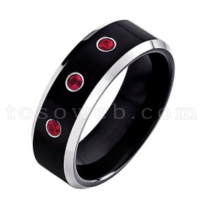 May include: A black and silver tungsten carbide ring with three red gemstones. The ring has a beveled edge and a polished finish.