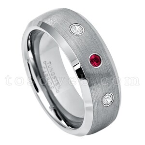 May include: A brushed silver tungsten carbide ring with a beveled edge and three gemstones. The ring has two small white gemstones and one larger red gemstone.