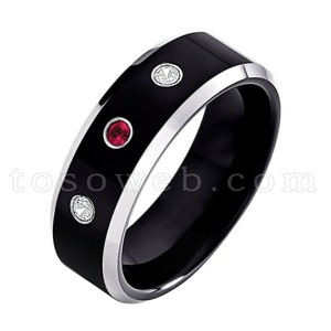 May include: A black ring with a silver-colored trim. The ring features three gemstones: two clear and one red. The ring has a glossy finish and a modern design.