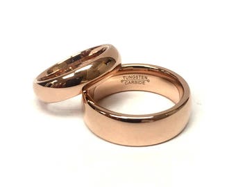 May include: Two rose gold tungsten carbide rings. One ring is slightly smaller and rests on top of the larger ring. The larger ring has the words "Tungsten Carbide" engraved on the inside. Both rings have a polished, reflective surface.