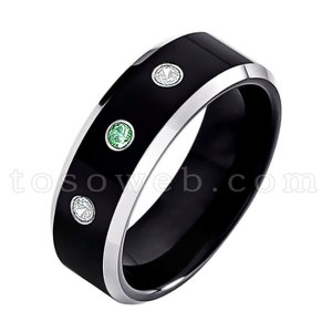 May include: A black ring with a silver trim and three gemstones. The gemstones are a mix of clear and green, set in a band of black. The ring has a modern, sleek design.