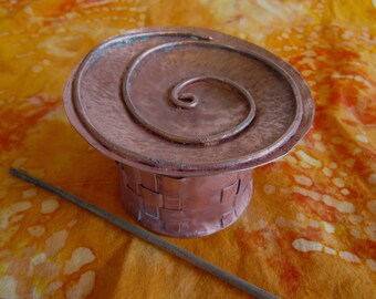 May include: A copper box with a woven pattern and a lid with a spiral design. The box is sitting on a patterned orange and yellow fabric.