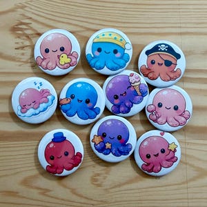May include: A collection of ten round, white buttons featuring cartoon octopuses in various colors and designs. Some octopuses wear hats, eye patches, or hold ice cream cones. The octopuses are pink, blue, purple, and red.