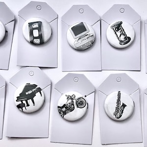 May include: A collection of black and white illustrated pinback buttons featuring various designs. The designs include a rotary phone, a VHS tape, a computer, an hourglass, a hand, a bicycle, a piano, a motorcycle, a saxophone, and a biplane.