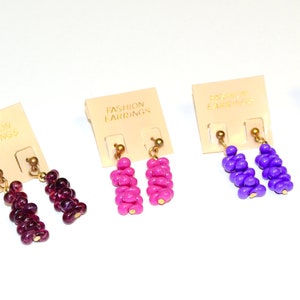 Pode incluir: Quatro pares de brincos pendurados nas cores vermelho escuro, rosa choque e roxo. Cada brinco apresenta um conjunto de contas suspensas por um gancho dourado. Os brincos são exibidos em cartões creme com a inscrição "FASHION EARRINGS".