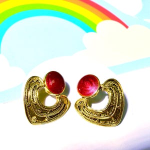 May include: A pair of gold-toned heart-shaped earrings with red cabochon accents. The earrings are set against a backdrop of a rainbow and clouds, creating a whimsical and colorful aesthetic.