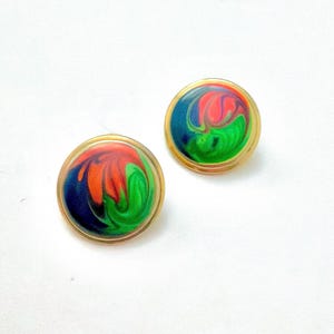 May include: A pair of circular stud earrings featuring a gold-toned metal rim surrounding a smooth, domed center. The surface displays a marbled, swirling pattern of vibrant orange, deep blue, and bright green colors blended together.