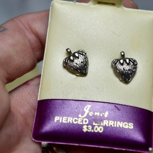 Pode incluir: Brincos perfurados em forma de morango prateados em um cartão creme com uma faixa roxa. O cartão diz "Jonet PIERCED EARRINGS". Os brincos têm superfícies texturizadas e pequenos caules.