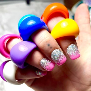 Puede incluir: Una mano muestra una colección de anillos coloridos y gruesos. Los anillos son en tonos rosa, morado, blanco, azul, naranja y amarillo. Las uñas están pintadas con purpurina y un diseño degradado rosa.