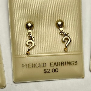 May include: Gold-coloured pierced earrings shaped like a question mark, presented on a cream card. The card is labelled "PIERCED EARRINGS". The earrings are attached to a small, round stud.