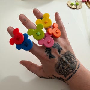 May include: A hand displays six colourful, Mickey Mouse-shaped buttons. The buttons are red, blue, green, yellow, orange, and pink. Each button has a contrasting thread sewn through it. The hand has tattoos of a tiger and the words "Calvin and Hobbes".