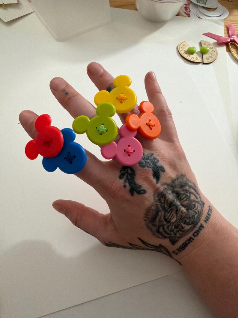 May include: A hand displays six colourful, Mickey Mouse-shaped buttons. The buttons are red, blue, green, yellow, orange, and pink. Each button has a contrasting thread sewn through it. The hand has tattoos of a tiger and the words "Calvin and Hobbes".