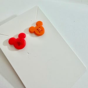 May include: Two Mickey Mouse-shaped buttons. One is bright red, the other is orange. Both have a cross-stitch design in the center. They are placed on a white card.