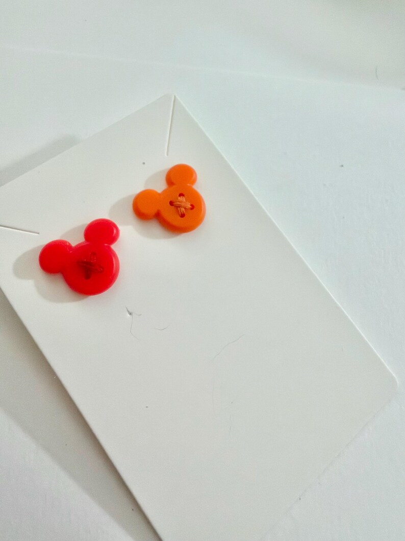 May include: Two Mickey Mouse-shaped buttons. One is bright red, the other is orange. Both have a cross-stitch design in the center. They are placed on a white card.