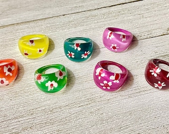 Op de afbeelding: Een verzameling van zeven dikke harsringen in verschillende kleuren, waaronder geel, oranje, groen, blauwgroen, roze en bordeaux. Elke ring is versierd met een wit bloemmotief met rode centra, wat een speels en kleurrijk accessoire creëert.